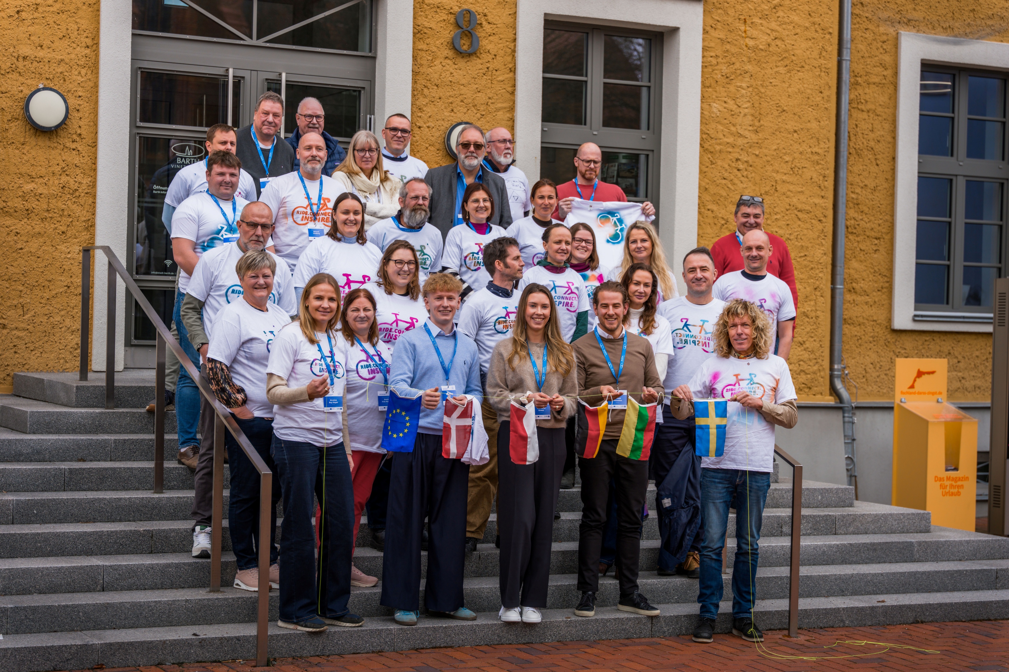 Eine Menschengruppe mit weißen T-shirts steht vor dem eingang eines Hauses (Vineta Bürgerhaus). die erste Reihe hält eine Kette aus kleinen Flaggen.